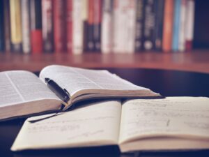 Two books open on a table in the foreground and a shelf full of books in the background.